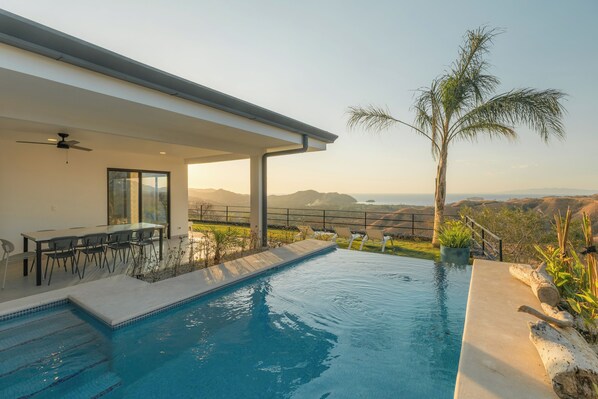 Una piscina con vistas a montañas, un porche cubierto con sillas y una palmera.