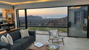 Ein modernes Wohnzimmer mit Sofa, Sessel und Couchtisch, einer Küchenzeile und einer großen Schiebeglastür, die auf einen Balkon mit Landschaftsblick führt.