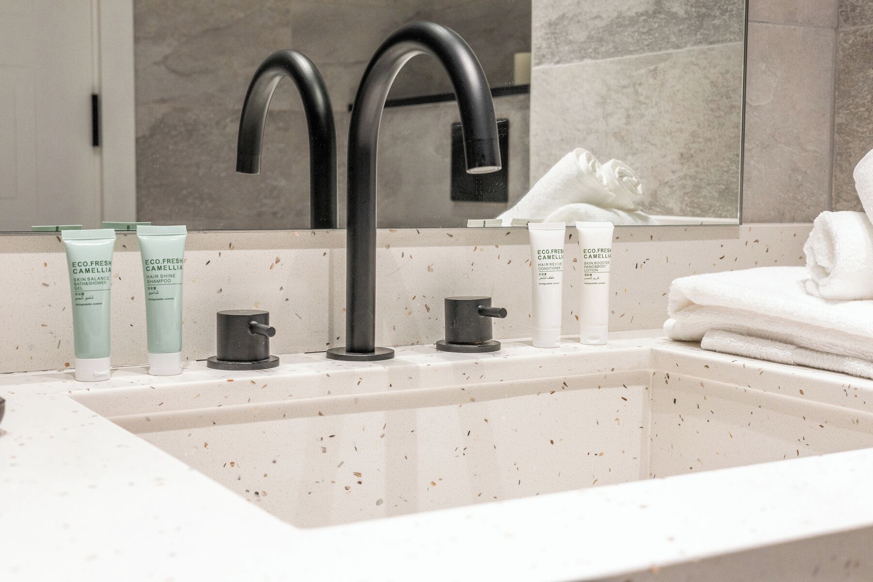 A bathroom with a white sink, two black faucets, and three bottles of Eco.Fresh Camellia hair care products.