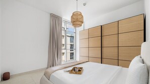 Ein modernes Schlafzimmer mit einem großen Bett, einem Fenster mit Vorhängen, einer Rattan-Pendelleuchte und Blick auf Gebäude.
