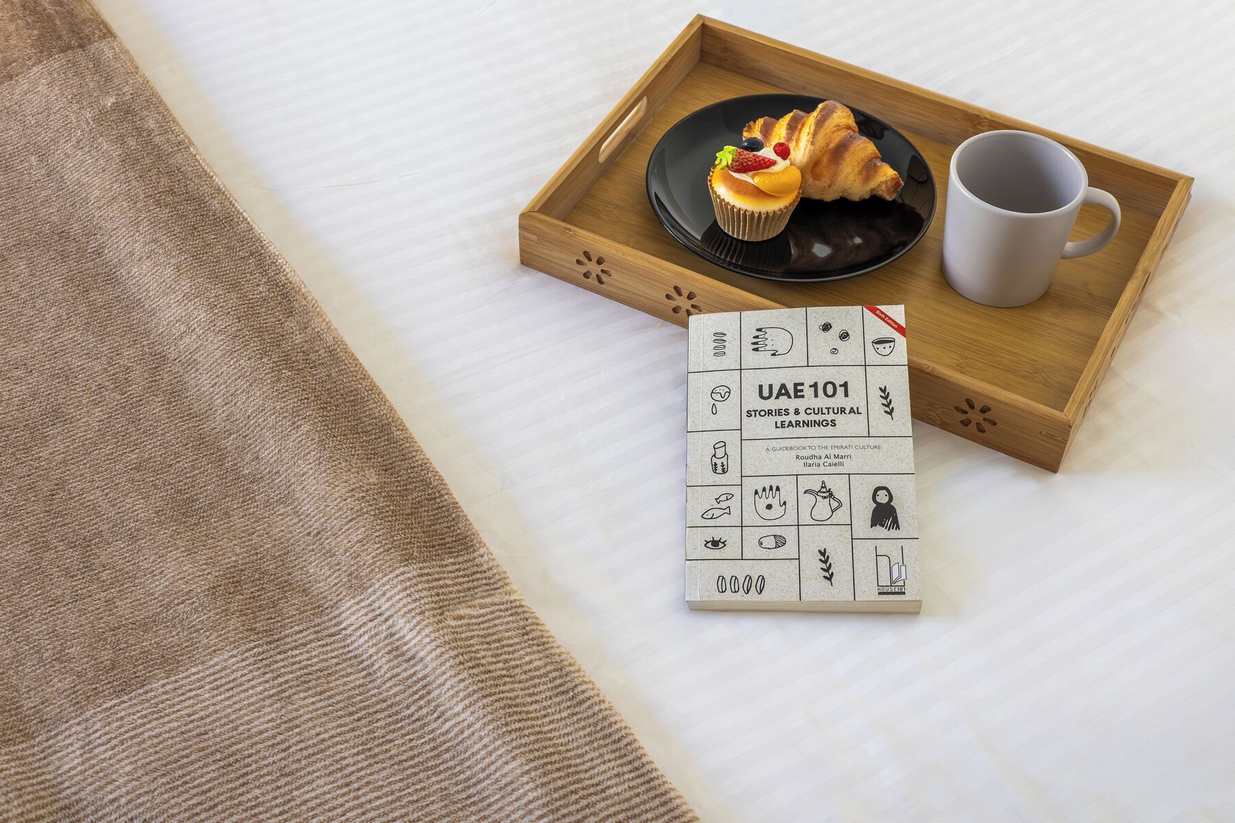 Ein Tablett mit einem Croissant, einem Cupcake und einer Tasse auf einem Bett mit einer beigen Decke.