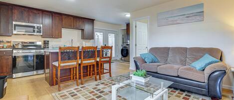 A modern kitchen with dark wood cabinets, a stainless steel microwave, and an oven. There is a dining area with a glass coffee table and a patterned rug. A living room with a gray sofa and a painting on the wall is visible.