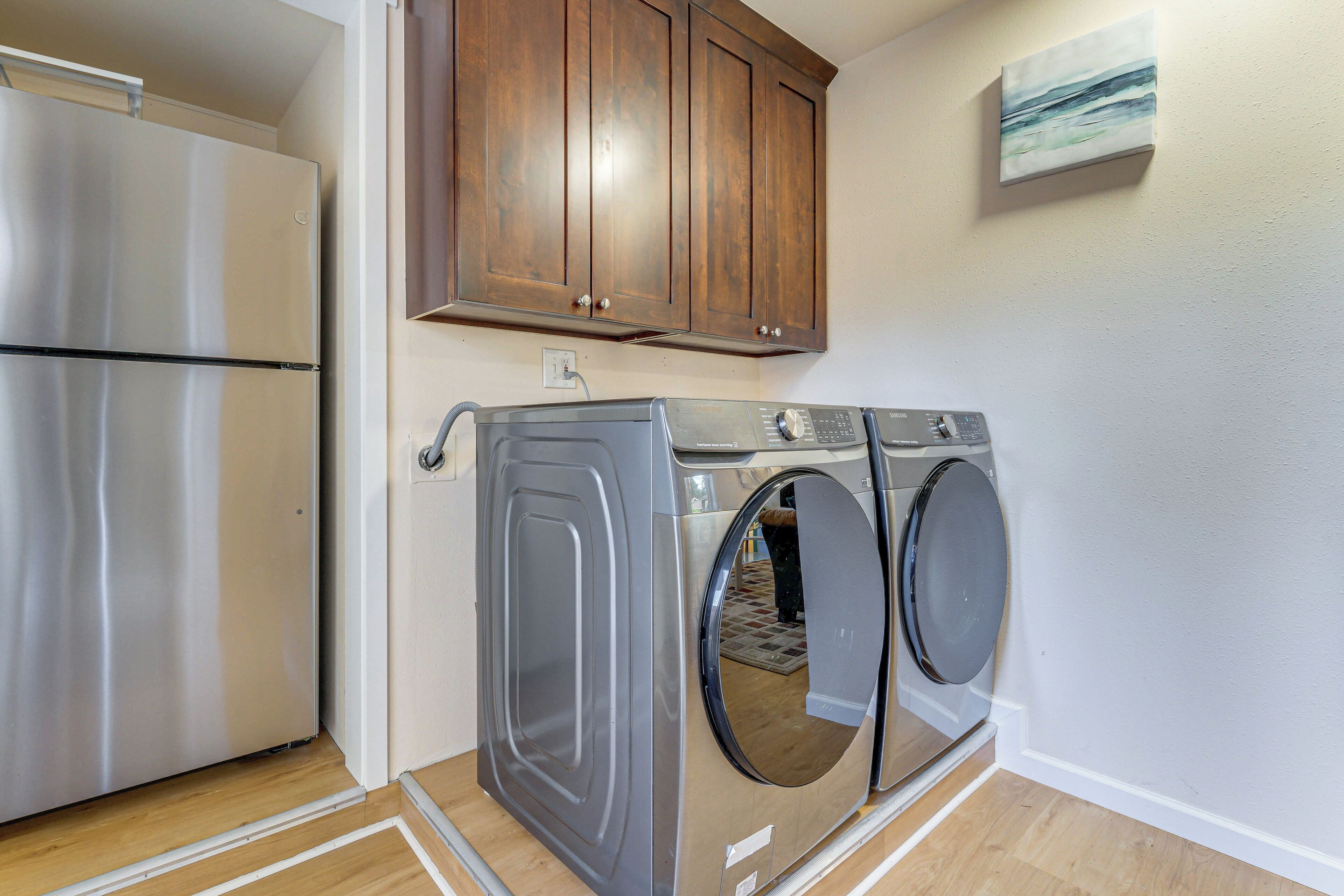 A compact laundry area with a stainless steel refrigerator, a gray washing machine, and wooden cabinets above.