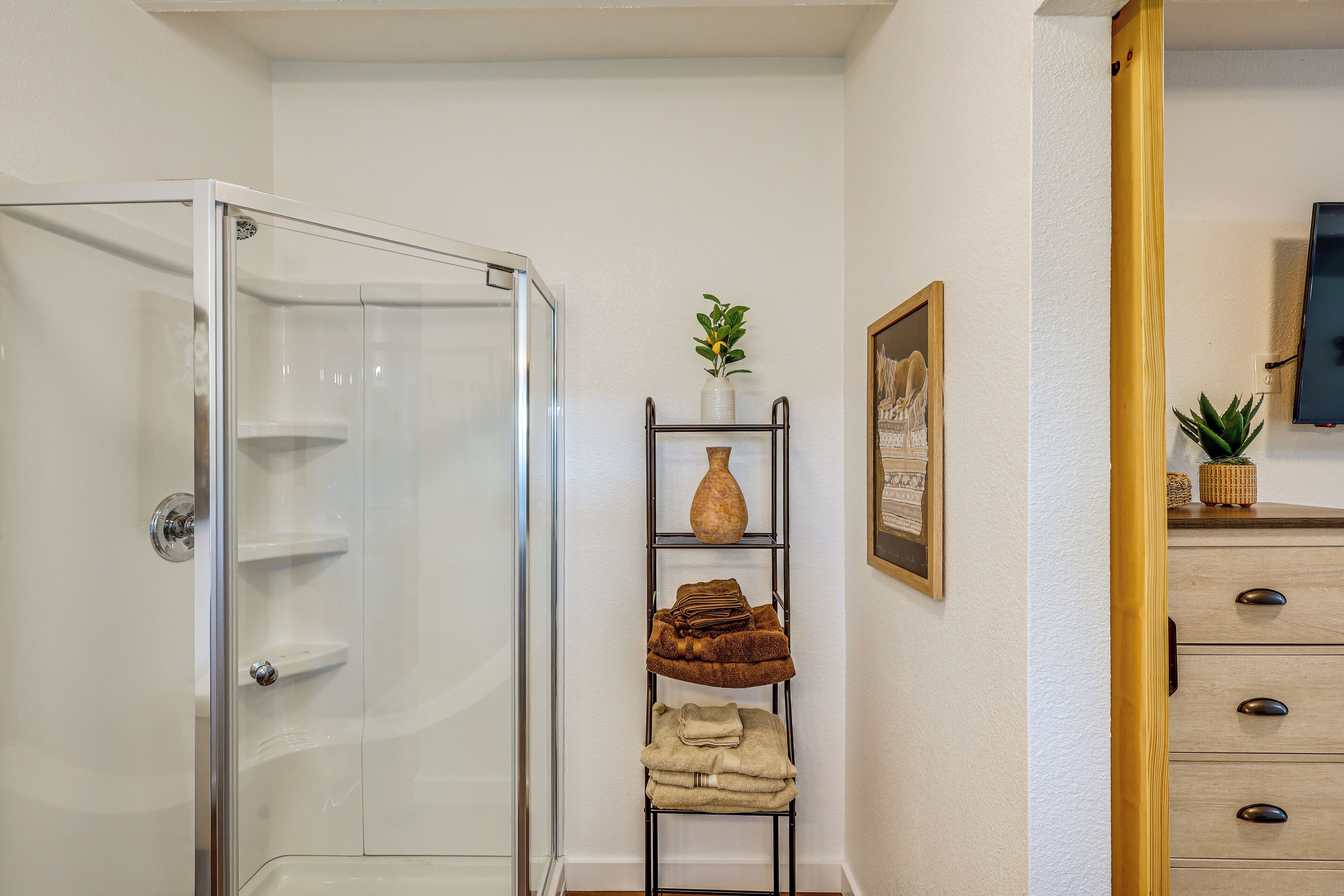 A bathroom with a glass shower enclosure, a towel rack with folded towels, a vase, and a framed picture on the wall.