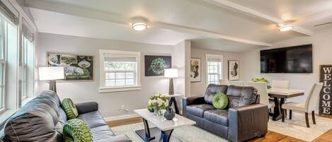 A modern living room with a gray sofa, a coffee table, and a dining area with a TV and a 'WELCOME' sign.