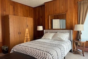 A bedroom with a wooden wardrobe, a bed with a patterned blanket, bedside lamps, and a mirror on the wall.