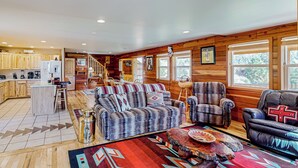 A spacious living area with a sofa, armchairs, and a coffee table. A kitchen is visible in the background with wooden cabinets and a refrigerator. - 3BR Rustic Cabin w/ 2 Decks & Hot Tub (Garden Valley)