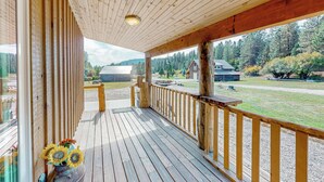 A wooden deck with a railing, a vase with sunflowers, and a view of a forested area with buildings in the background. - 3BR Rustic Cabin w/ 2 Decks & Hot Tub (Garden Valley)