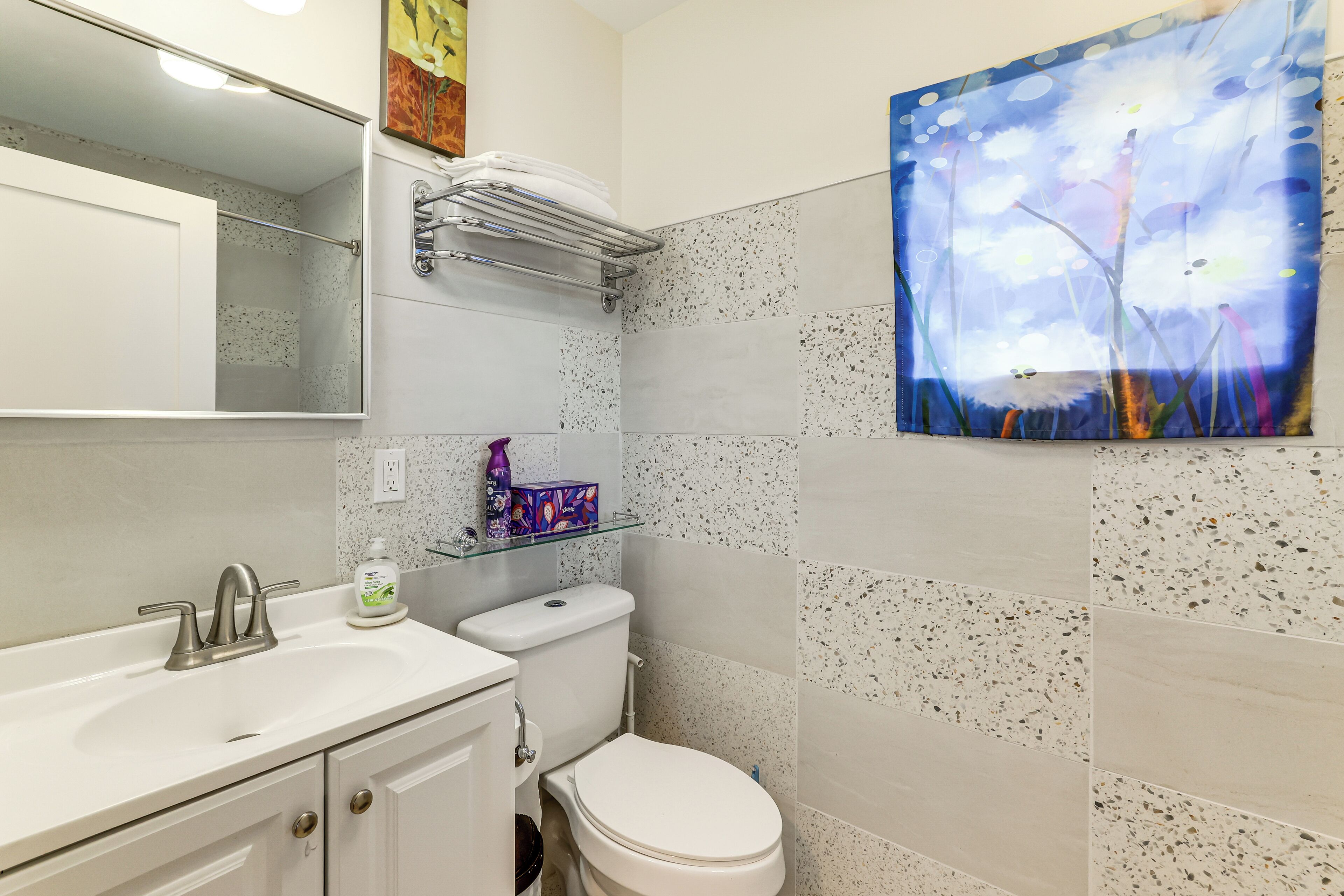 A bathroom with a white sink, a toilet, and a shower. There is a painting on the wall and a shelf with toiletries.