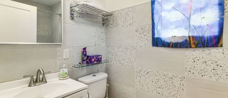A bathroom with a white sink, a toilet, and a shower. There is a painting on the wall and a shelf with toiletries.