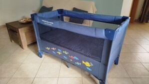 Berço azul com cobertura de malha e adesivos de peixe, posicionado em um quarto com piso de azulejos e uma cama ao fundo.