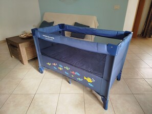 A blue baby crib with a mesh top and fish decals, positioned in a room with a tiled floor and a bed in the background. - ONDA DO MAR. Cozy apartment near the sea in Santa María (Santa Maria)