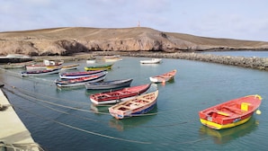 Um porto com várias embarcações menores atracadas, um farol ao fundo e um litoral rochoso.