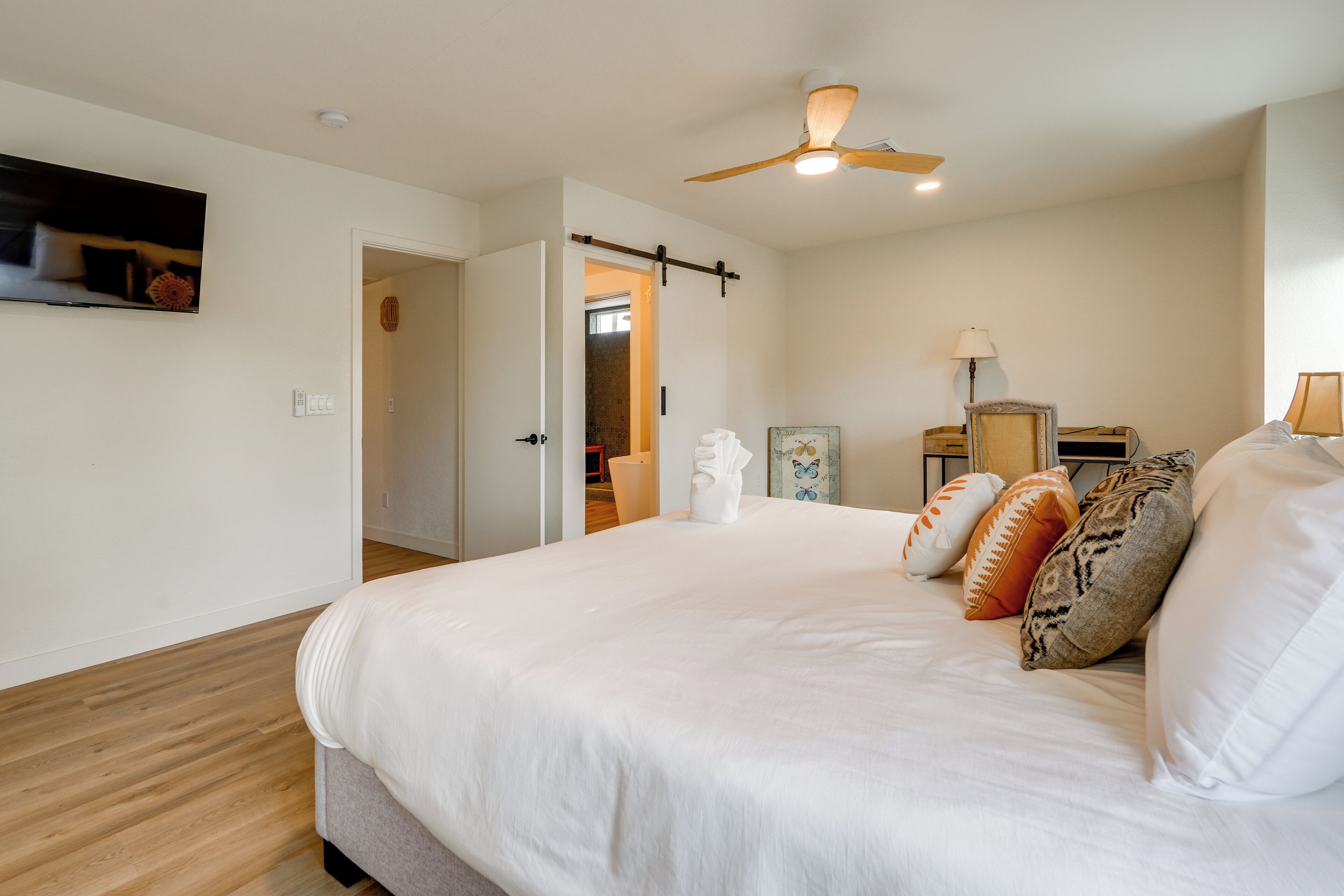 A bedroom with a large bed, a ceiling fan, a TV mounted on the wall, and a door leading to another room.