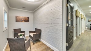 A hallway with a white brick wall, a black door with a decorative handle, and a dining area with wicker furniture and a painting. - Walk to Park: Modern Apt 2 Mi to Union College! (Schenectady)
