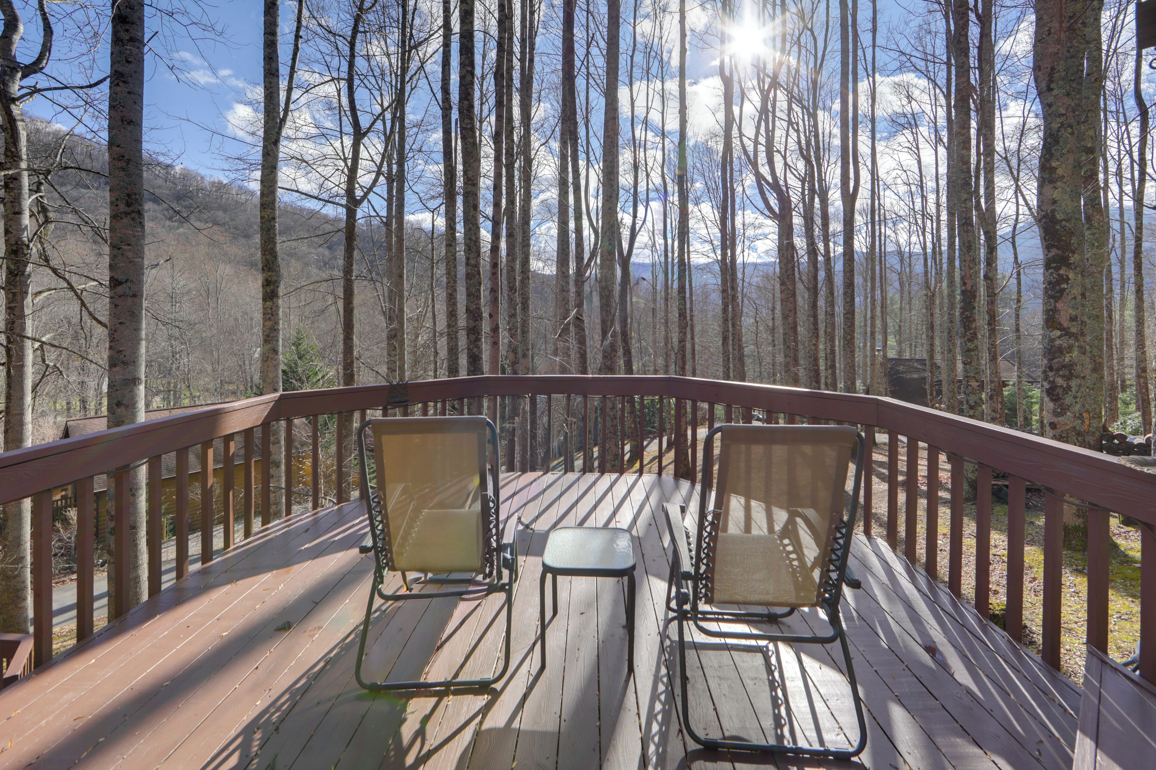 A wooden deck with two metal chairs and a small table, surrounded by tall trees and a clear sky.