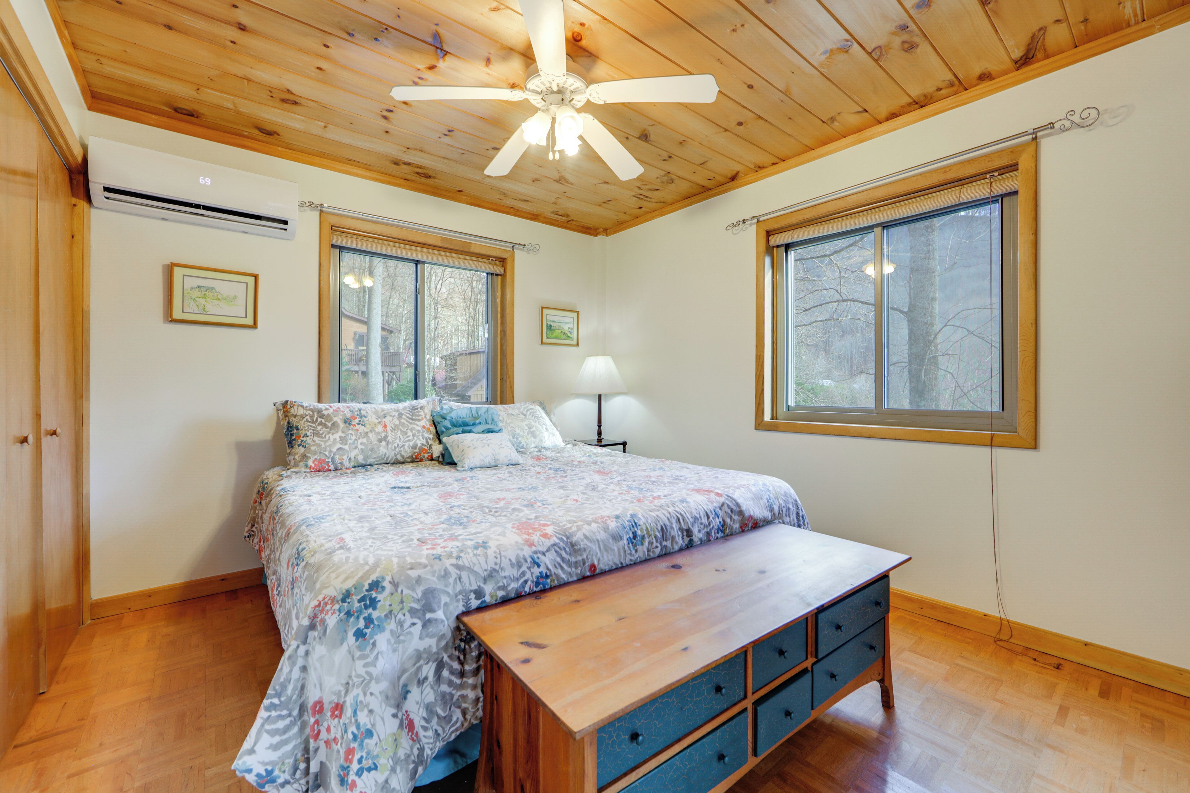 A bedroom with a wooden ceiling, large windows, a bed with a floral bedspread, a bedside table, and a bench.