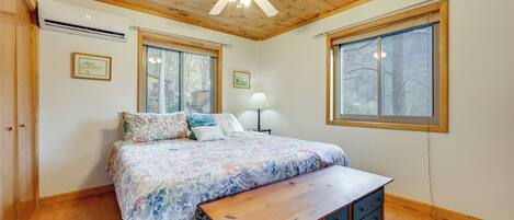 A bedroom with a wooden ceiling, large windows, a bed with a floral bedspread, a bedside table, and a bench.