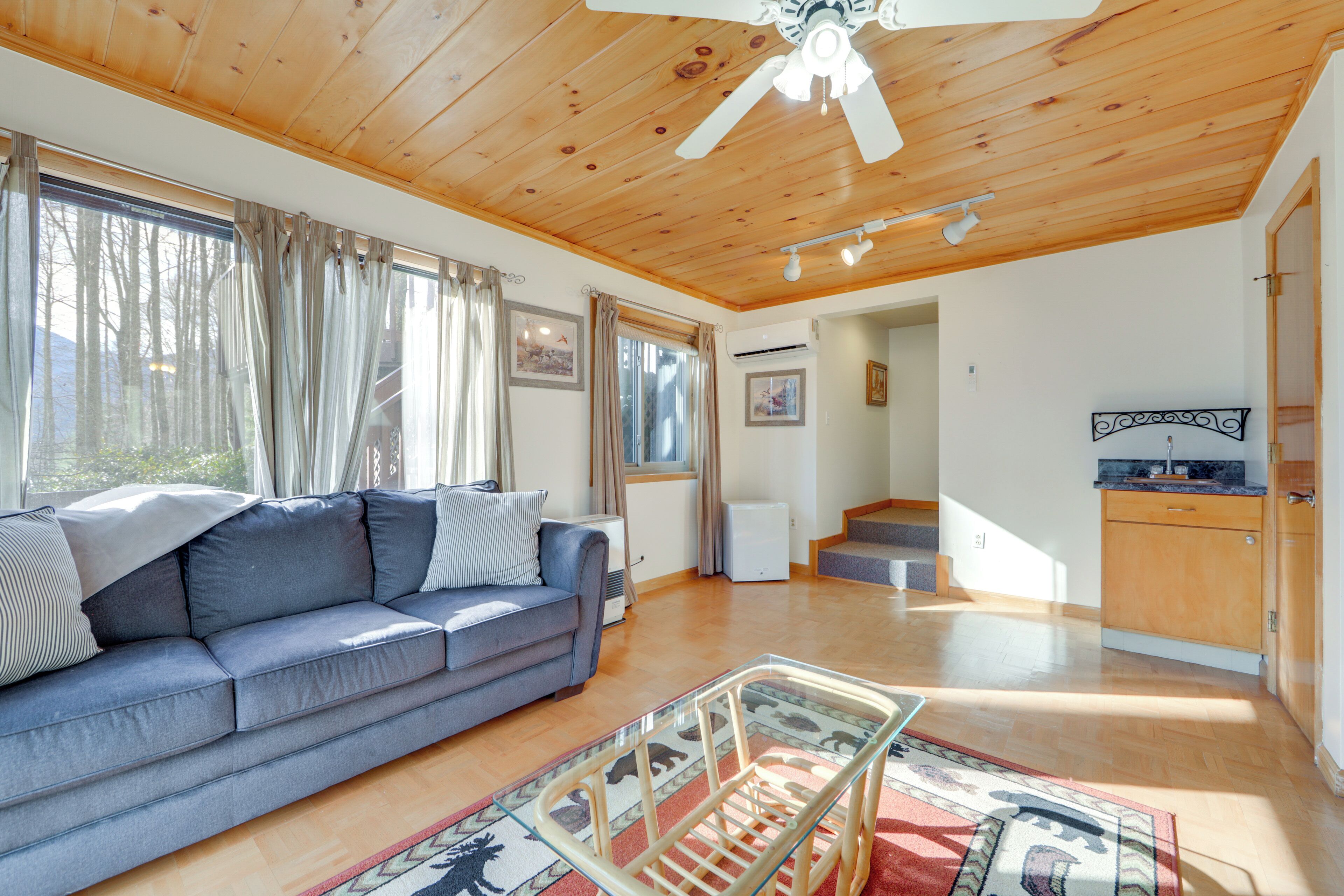 A living room with a wooden ceiling, a sofa, a glass coffee table, and a kitchen area with wooden cabinets.