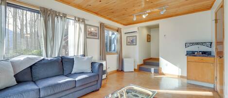 A living room with a wooden ceiling, a sofa, a glass coffee table, and a kitchen area with wooden cabinets.