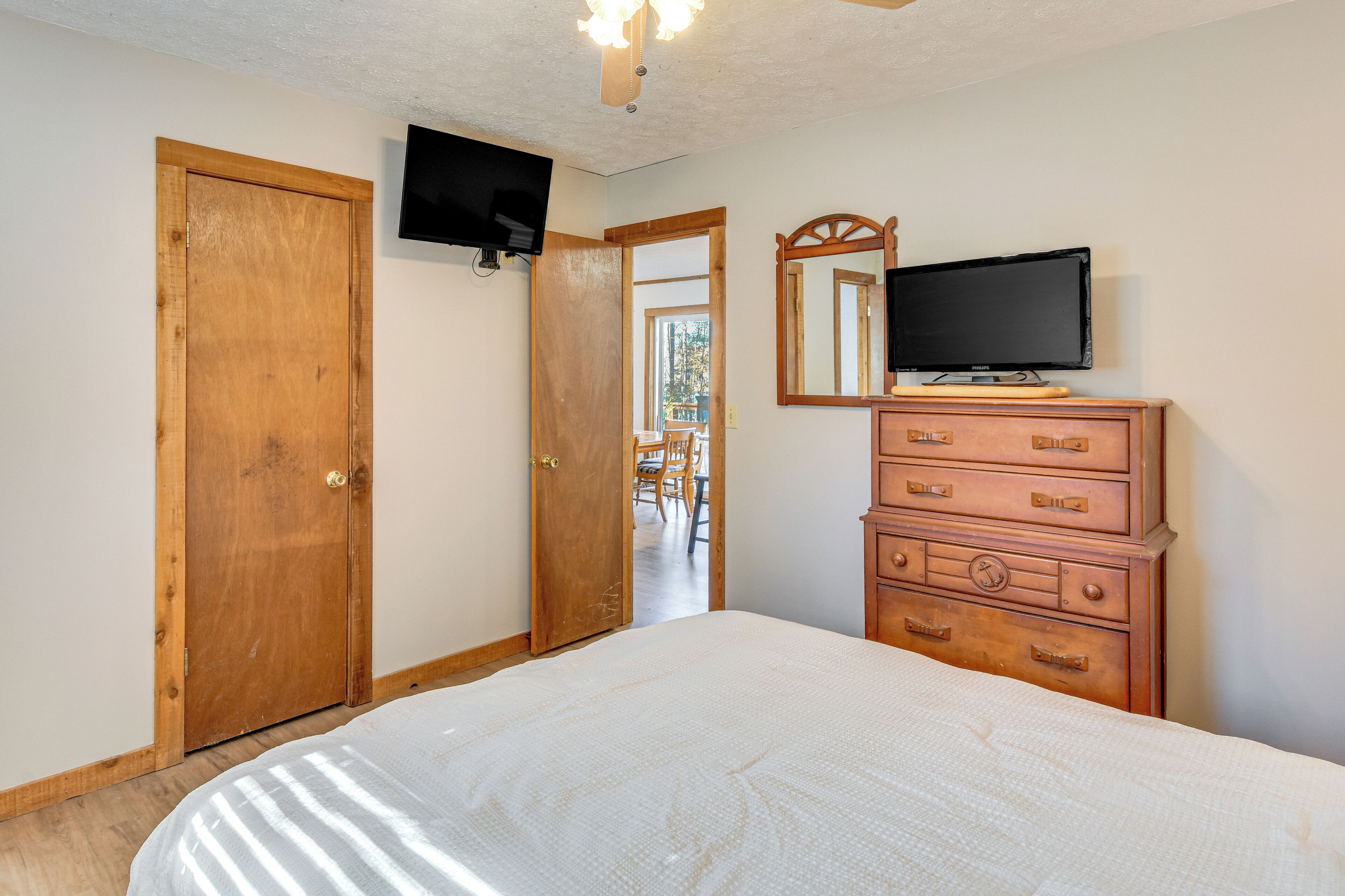 A bedroom with a bed, wooden dresser, two wooden doors, a television mounted on the wall, and a mirror above the dresser.