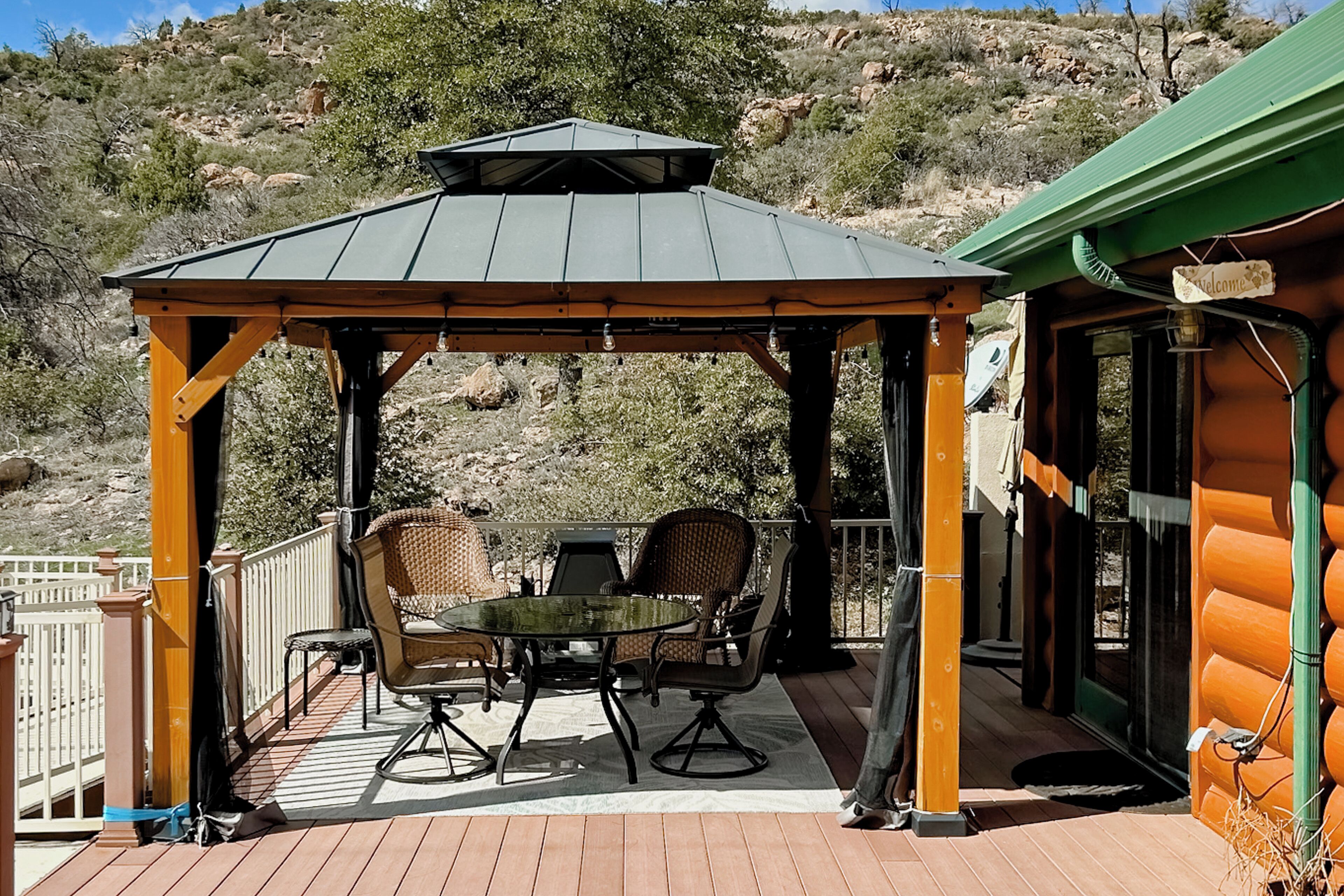 Un espace extérieur aménagé, abrité et doté d’une table et de chaises, d’une pergola, et offrant une vue sur un paysage vallonné.