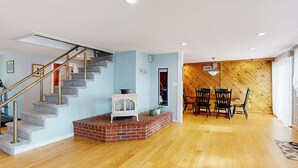 Un vaste espace de vie doté d’un plancher en bois, d’une cheminée en briques, d’un escalier avec une rampe en verre et d’un coin repas avec des chaises et une table en bois.