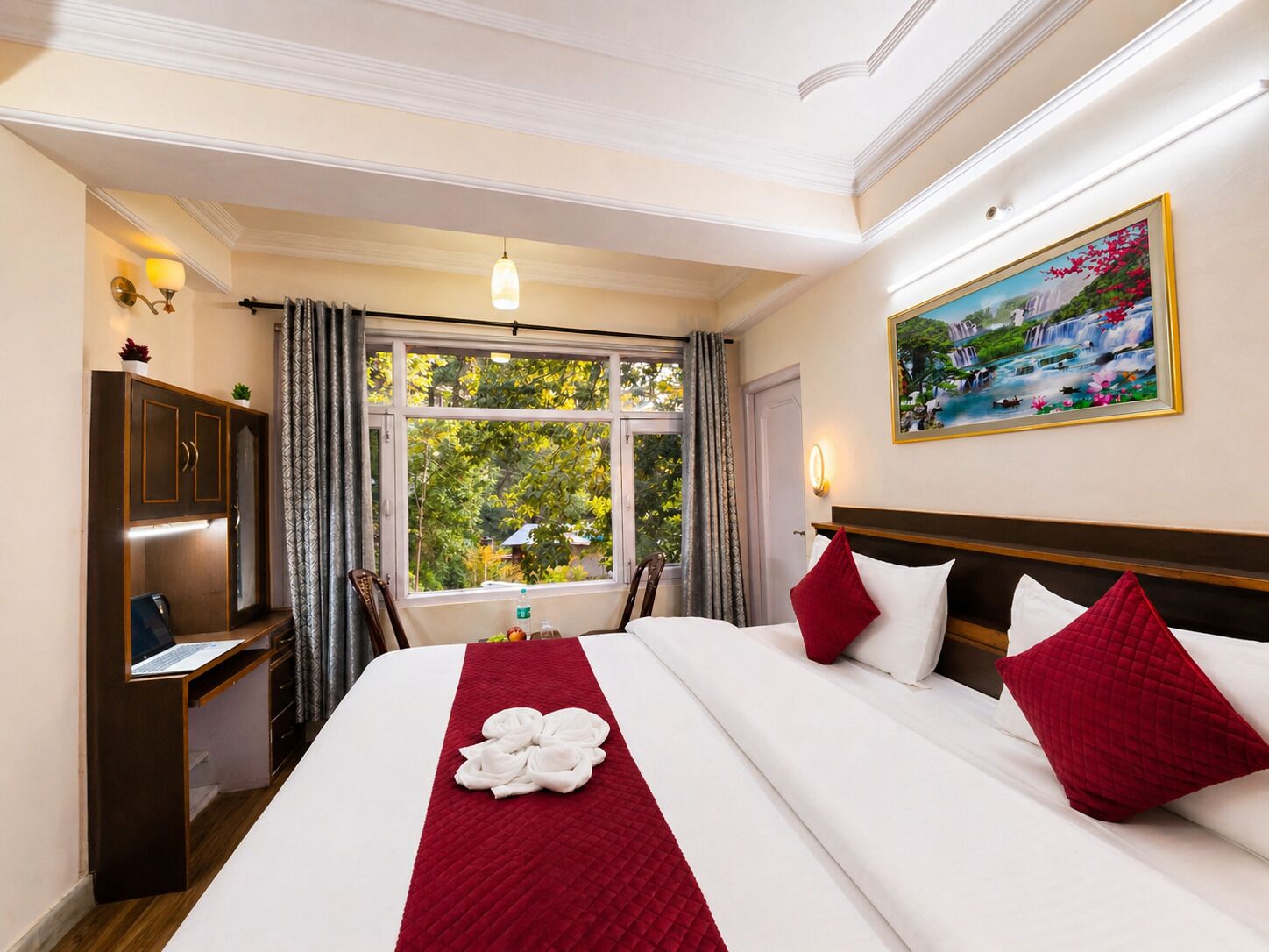 A hotel room with a bed, a desk, a wardrobe, a window with a view of greenery, and a painting on the wall.