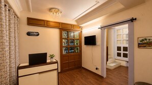 Ein Zimmer mit Schreibtisch, Fernseher, einer Kristallleuchtre, einer Glastür und einer kleinen Pflanze.