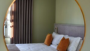 A circular mirror reflecting a bed with white linens and two brown pillows, a gray headboard, and a window with blinds. - Megaro Gioule (Voio)