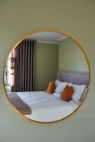 Un espejo circular refleja una cama con sábanas blancas y dos almohadas marrones, un cabecero gris y una ventana con persianas.