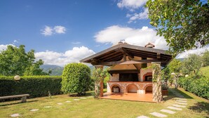 Eine Außenküche aus Stein mit Pergola, umgeben von einem gepflegten Garten und freiem Blick auf die Berge.