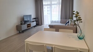 A modern living room with a flat-screen TV, a sofa, a dining table with chairs, and a small plant.