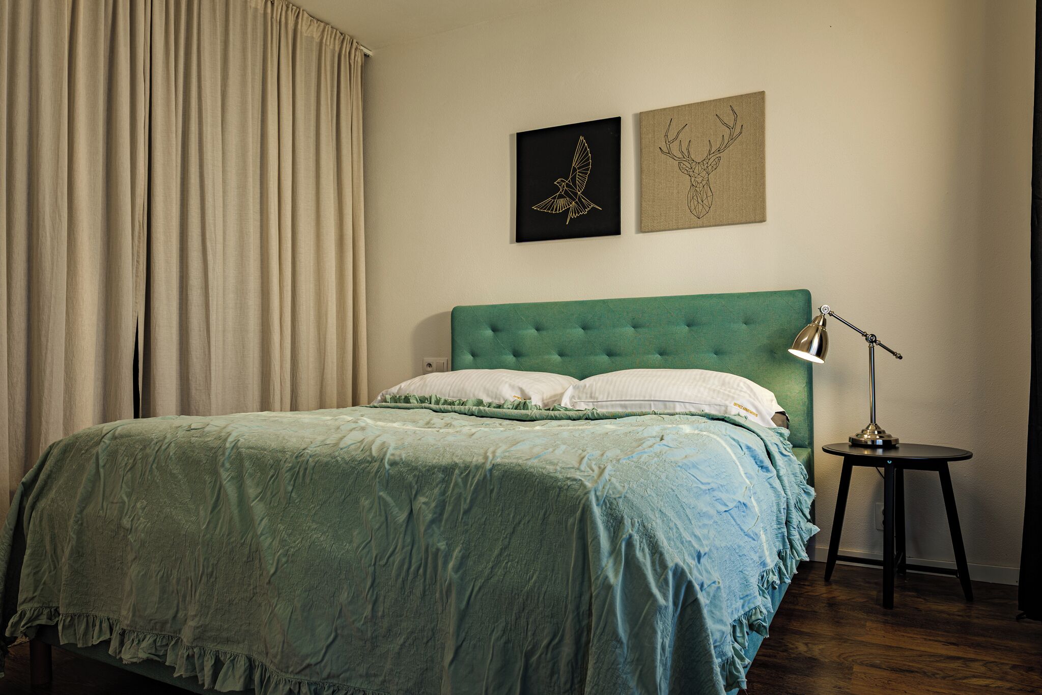 A bedroom with a green upholstered headboard, a bed with white pillows, a bedside lamp, and two framed artworks on the wall.