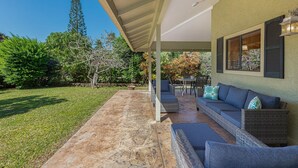 A covered patio area with outdoor furniture, a stone floor, and a view of a garden with trees and shrubs. - Hale Hauoli - lovely home, near golf & beaches (Princeville)