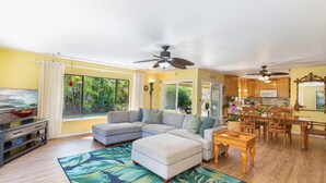 A spacious living room with a large sectional sofa, a wooden dining table, and a kitchen area with wooden cabinets and a microwave. - Hale Hauoli - lovely home, near golf & beaches (Princeville)
