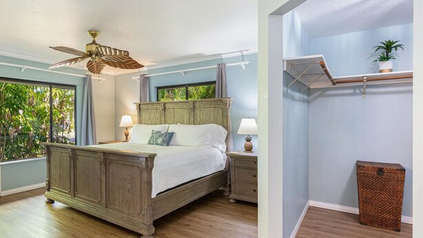 A bedroom with a wooden bed, a ceiling fan, a nightstand, a lamp, a small table, a plant, and a window with a view of greenery. - Hale Hauoli - lovely home, near golf & beaches (Princeville)