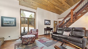 A spacious living room with a wooden ceiling, a brown leather sofa, a patterned ottoman, and a large window offering a view of trees. - 2 Miles to Deadwood! 3.5 Acres w/ 4,100 Sq. Ft - Hot Tub, Paved! (Sturgis)