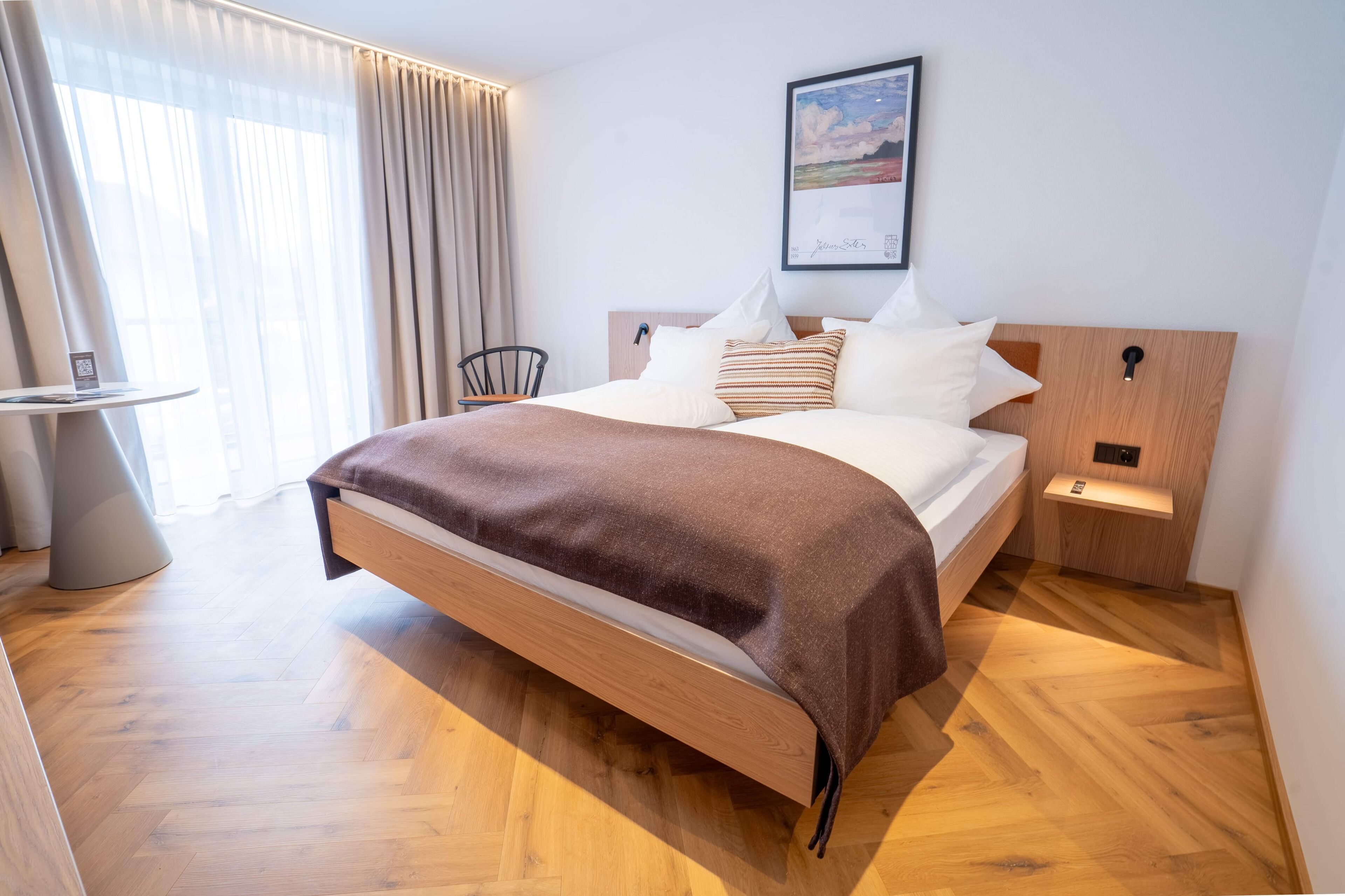 A modern hotel room with a wooden bed, a bedside table, a chair, a small table, and a painting on the wall.