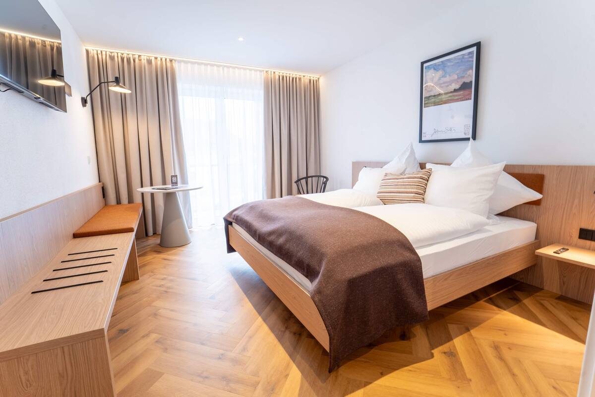 A modern hotel room with a wooden bed, a bench, a small table, and a wall-mounted TV.