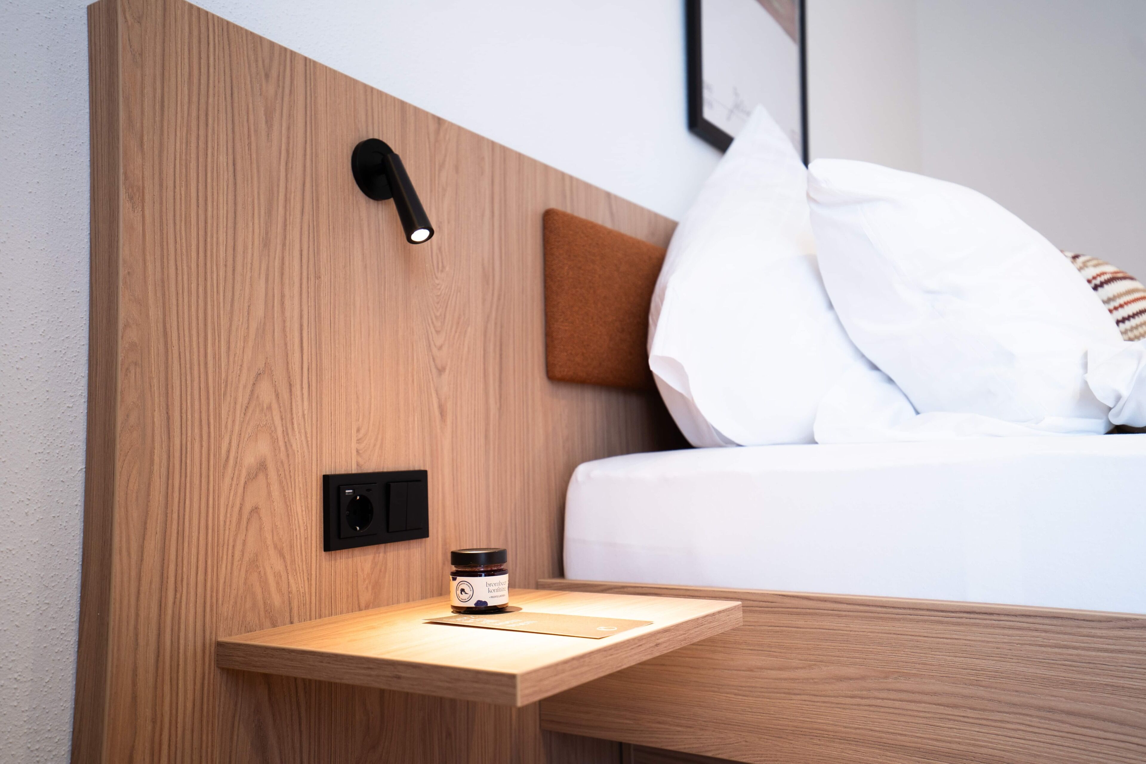 Un lit bien fait, avec du linge de lit blanc et un oreiller marron, une table de chevet sur laquelle se trouve un petit récipient, et un luminaire fixé au mur.