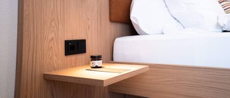 Un lit bien fait, avec du linge de lit blanc et un oreiller marron, une table de chevet sur laquelle se trouve un petit récipient, et un luminaire fixé au mur.