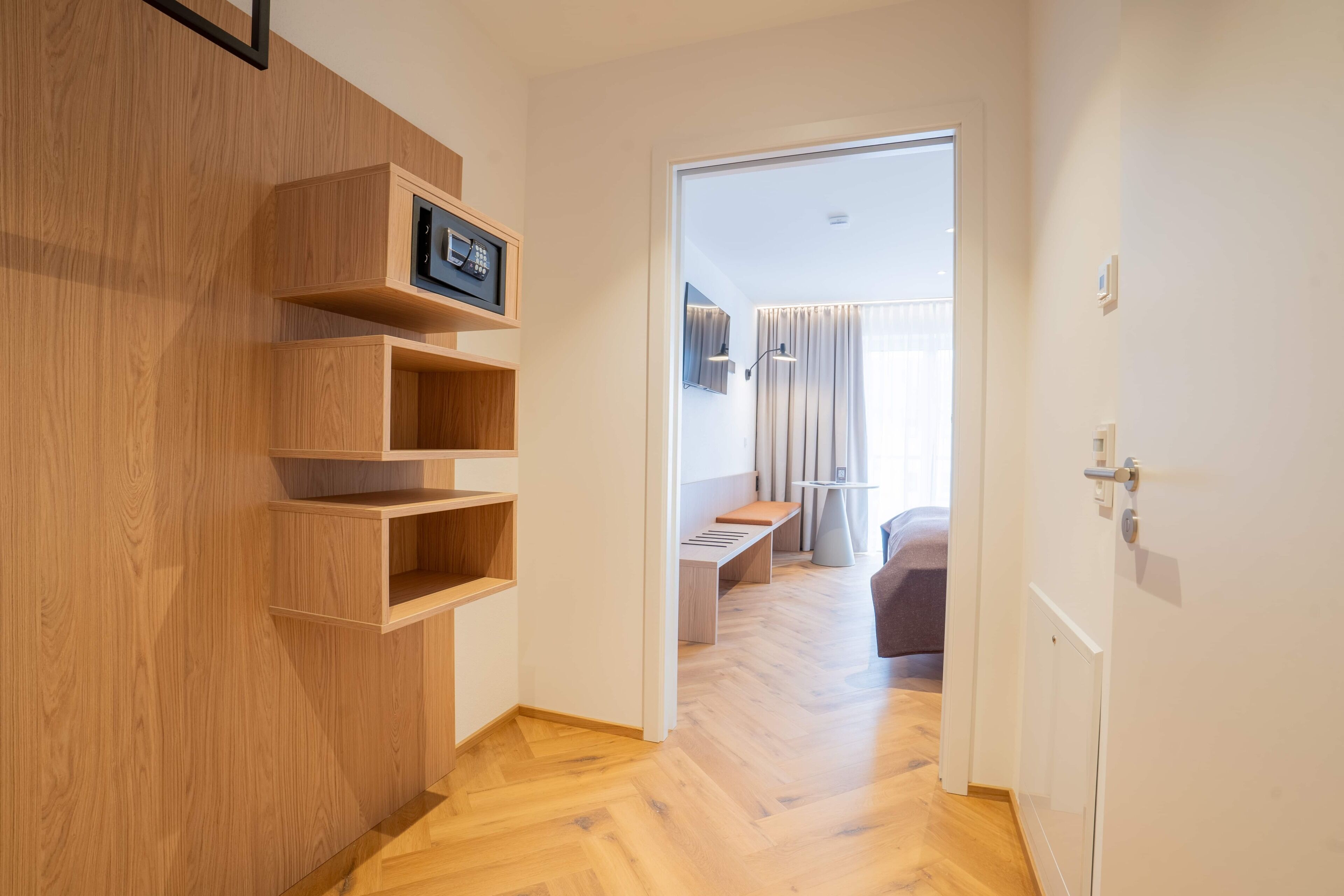A hallway with wooden shelving, a bench, and a door leading to a room with a bed and curtains.