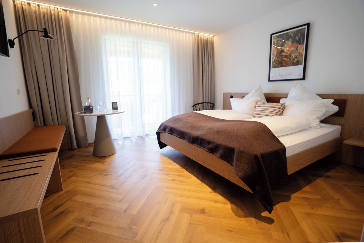 A hotel room with a wooden bed, a bedside table, a bench, and a large window with curtains.