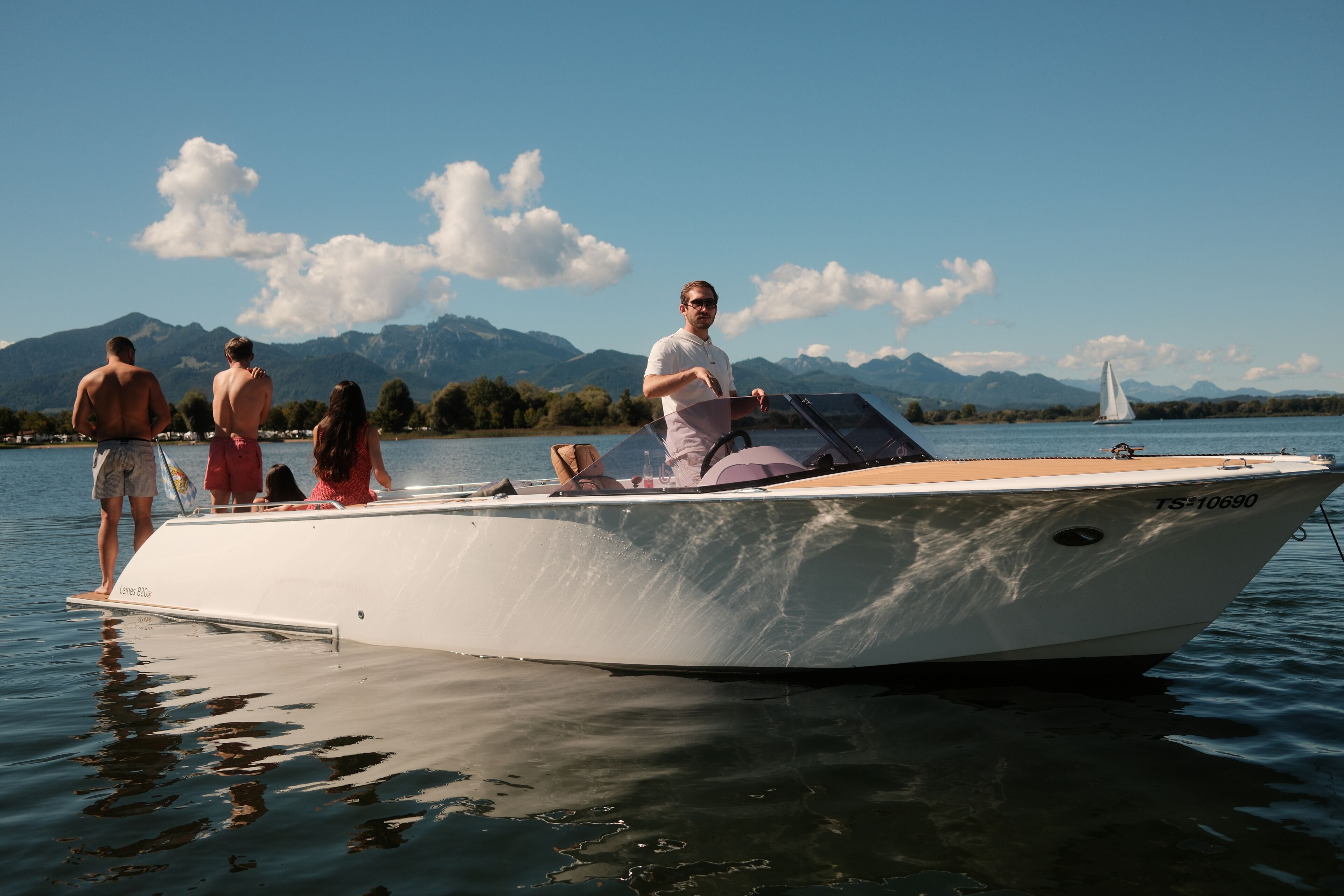 A boat with a person steering, passengers on board, and a mountainous landscape in the background.