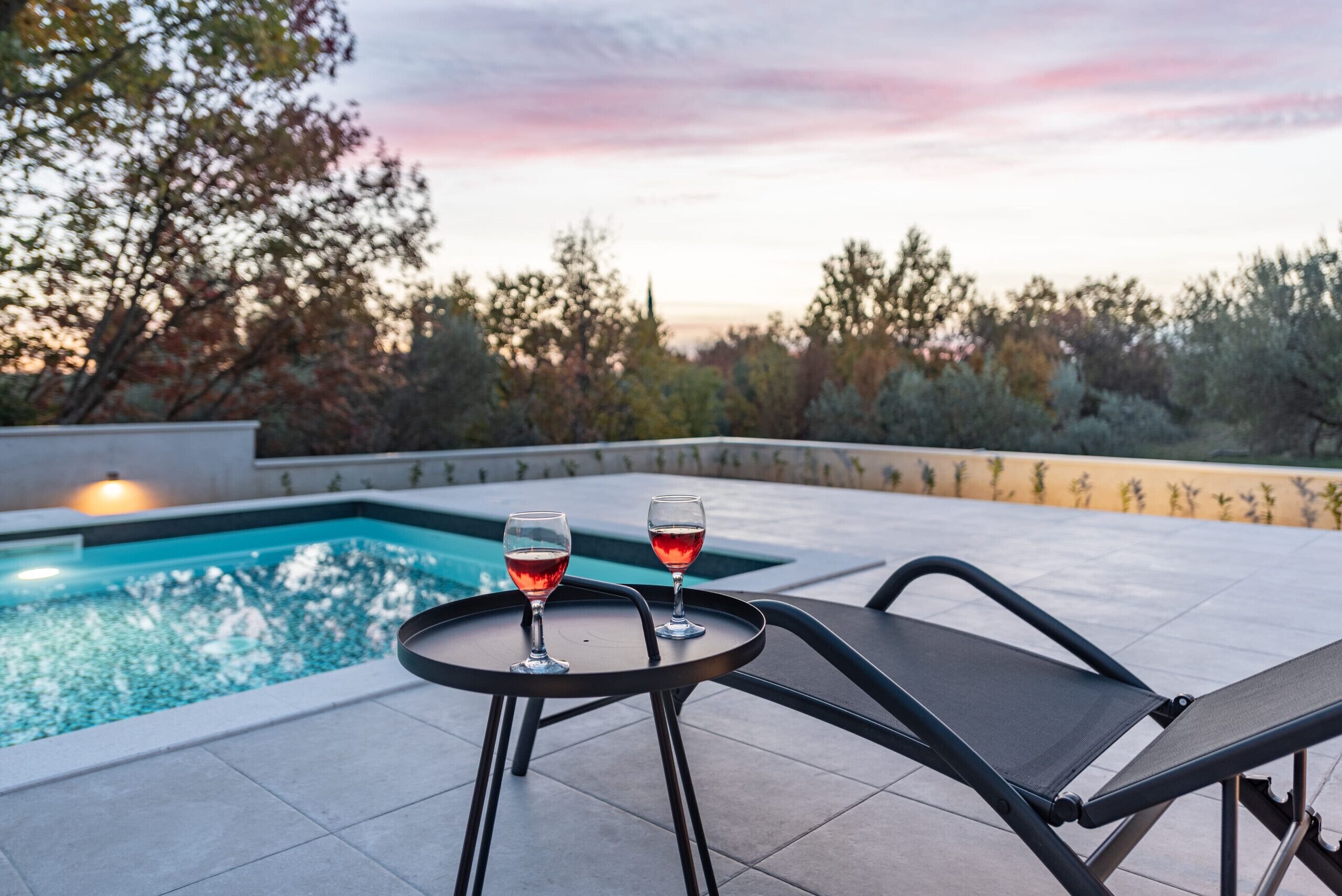 A poolside area with a lounge chair and two glasses of red wine on a small table.