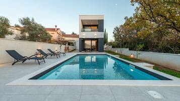 Une maison moderne à deux étages, dotée d’une piscine, de chaises longues et d’un jardin bien entretenu.