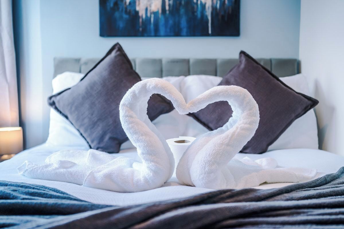 A bed with a heart-shaped towel arrangement, a grey headboard, and a painting on the wall.