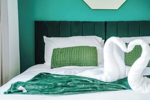 Un lit aux couleurs vert et blanc, avec une tête de lit sombre et une serviette pliée en forme de cœur.