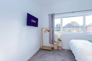 A bedroom with a bed, a TV mounted on the wall, a window with a view of houses, and a small bedside table with a lamp.